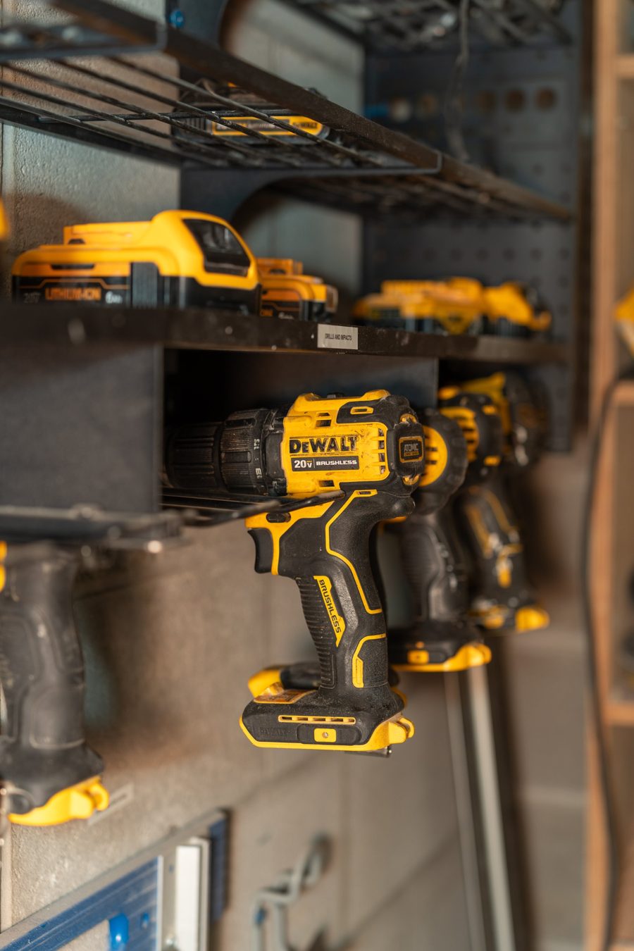 A row of yellow cordless drills on a wall rack with batteries on the shelf above