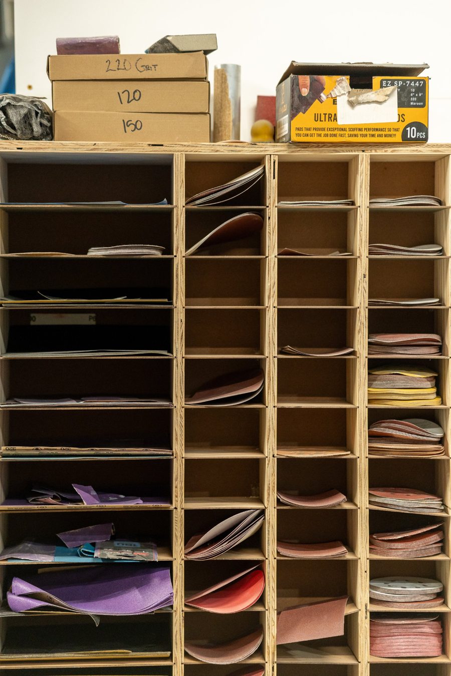 A plywood cubby shelf holding sheets and discs of sandpaper sorted by grit