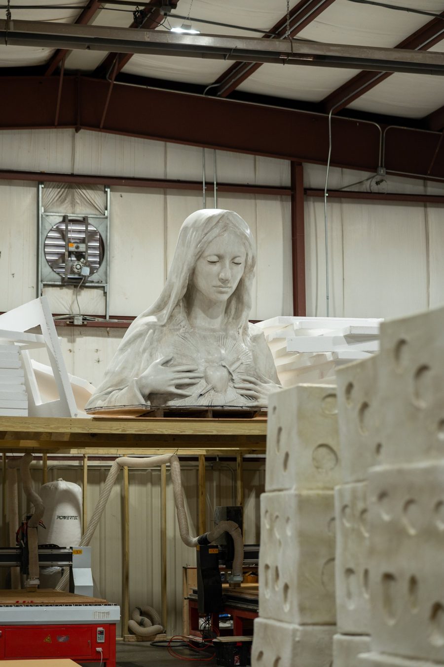 A large carved sculpture bust on a loft above CNC routers, with carved foam blocks in the foreground