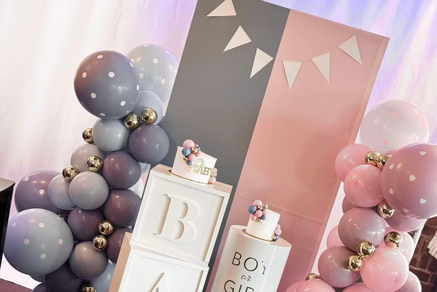 A gender-reveal backdrop with pink and gray panels, baby blocks and balloon garlands