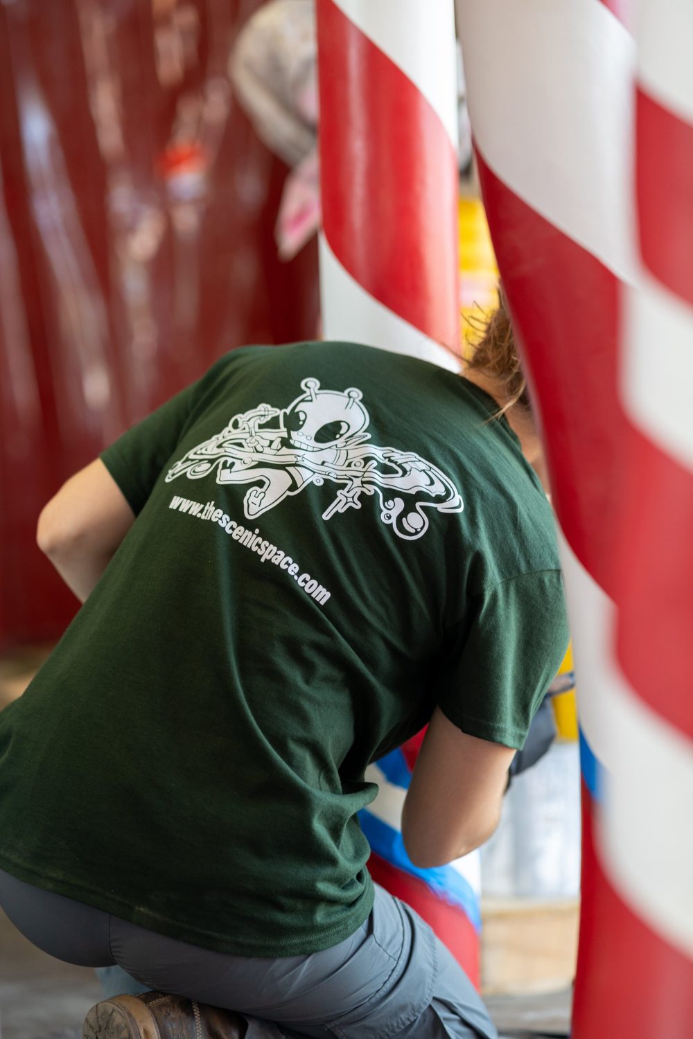 A Scenic Space painter, alien mascot on the back of their shirt, painting red and white striped columns